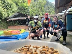 Three kayakers in helmets and PFDs pose next to a tray of grilled chonta curo grubs, with colorful kayaks and lush green forest in the background.